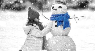 Ein kleines Mädchen mit Winterjacke und Mütze baut im Schneefall einen Schneemann. Der Schneemann trägt einen leuchtend blauen Schal und eine Mütze.
