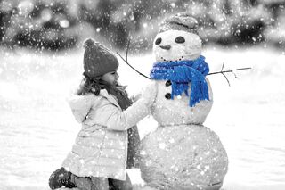 Ein kleines Mädchen mit Winterjacke und Mütze baut im Schneefall einen Schneemann. Der Schneemann trägt einen leuchtend blauen Schal und eine Mütze.